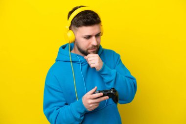 Sarı arka planda izole edilmiş bir bilgisayar oyunu kontrolörüyle oynayan genç beyaz adam yana bakıyor ve gülümsüyor.