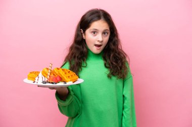 Pembe arka planda yüzünde şaşırmış ve şok olmuş bir yüz ifadesiyle waffle tutan beyaz bir kız.