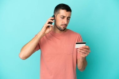 Genç beyaz adam mavi arka planda izole edilmiş cep telefonuyla kredi kartıyla alışveriş yapıyor.