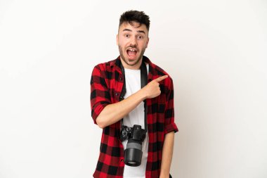 Beyaz arka planda izole edilmiş genç fotoğrafçı şaşırmış ve işaret ediyor.
