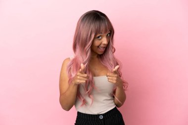 Pembe saçlı genç melez kadın pembe arka planda izole edilmiş şaşırmış ve ön tarafı işaret ediyor.