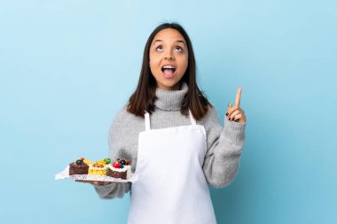 Pasta şefi büyük bir pasta tutuyor. Mavi arka planda. Şaşırmış ve işaret ediyor..