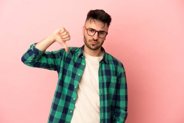 Pembe arka planda izole edilmiş genç beyaz adam olumsuz ifadeyle baş parmağını aşağı gösteriyor.