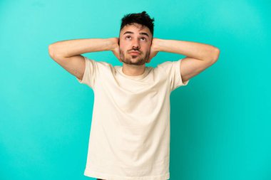 Mavi arka planda izole edilmiş genç beyaz adam hayal kırıklığına uğramış ve kulaklarını kapatmış.