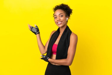 Sarı arka planda izole edilmiş genç Afrikalı spor kadını davet etmek için ellerini yana uzatıyor.