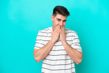 Kafkasyalı genç adam mavi arka planda izole edilmiş mutlu ve gülümseyen elleriyle ağzını kapatıyor.