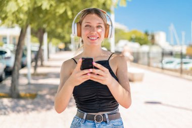 Genç sarışın kadın açık havada müzik dinliyor cep telefonu ve ön tarafa bakıyor.