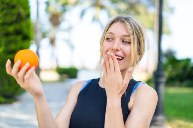 Sarışın, sporcu bir kadın elinde turuncu bir şeyle dışarıda bekliyor. Şaşırmış ve şok olmuş yüz ifadesi var.