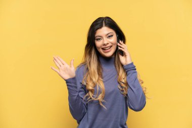 Sarı arka planda izole edilmiş cep telefonu kullanan genç Rus kızı mutlu bir ifadeyle selamlıyor.