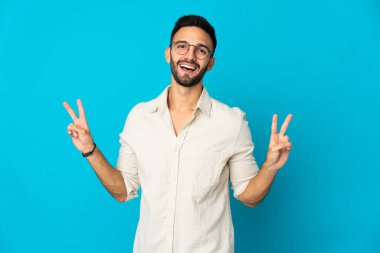 Mavi arka planda izole edilmiş genç beyaz adam iki eliyle zafer işareti gösteriyor