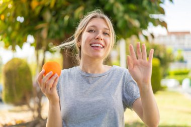 Dışarıda elinde portakal tutan genç sarışın kadın mutlu bir ifadeyle selamlıyor.