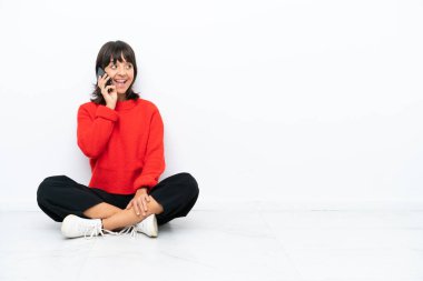 Genç melez kadın, beyaz arka planda izole bir şekilde yerde oturuyor ve cep telefonuyla sohbet ediyor.