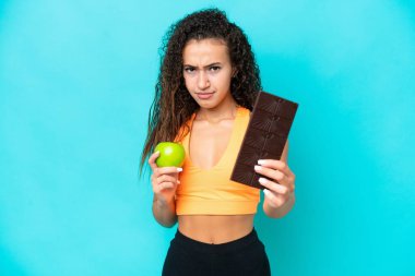 Genç bir Arap kadın mavi arka planda izole edilmiş. Bir elinde çikolata tableti, diğerinde elma alıyor.