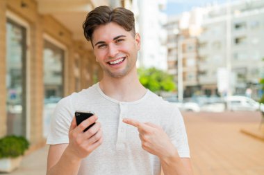 Genç yakışıklı adam cep telefonu kullanıyor ve onu doğrultuyor.