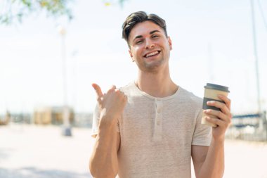 Genç ve yakışıklı bir adam açık havada kahvesini yudumlarken yan tarafı göstererek bir ürün sunuyor.