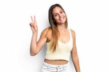 Beyaz arka planda izole edilmiş genç beyaz kadın gülümsüyor ve zafer işareti gösteriyor.