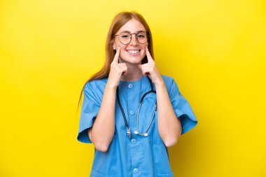 Sarı arka planda izole edilmiş genç kızıl saçlı hemşire mutlu ve hoş bir ifadeyle gülümsüyor.