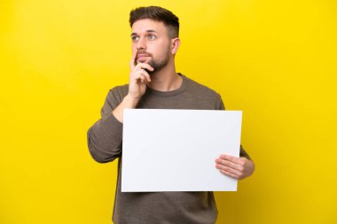 Kafkasyalı genç adam sarı arka planda izole edilmiş boş bir levha tutuyor ve düşünüyor.