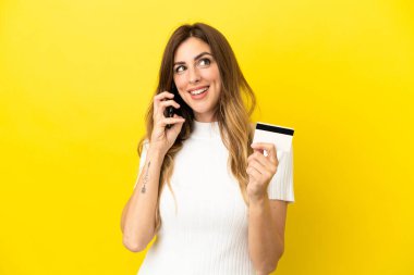 Beyaz kadın sarı arka planda izole edilmiş cep telefonuyla sohbet ediyor ve elinde bir kredi kartı tutuyor.