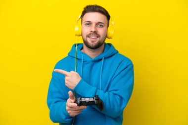 Sarı arka planda izole edilmiş bir bilgisayar oyunu kontrolörüyle oynayan genç beyaz adam bir ürünü sunmak için kenarı işaret ediyor.