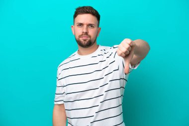 Mavi arka planda izole edilmiş genç beyaz adam olumsuz ifadeyle baş parmağını aşağı gösteriyor.