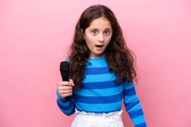Küçük şarkıcı kız pembe arka planda izole edilmiş bir mikrofonu kaldırıyor. Şaşırmış ve şok olmuş bir yüz ifadesi var.