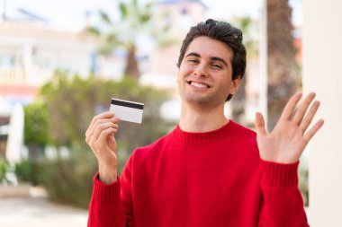 Genç ve yakışıklı bir adam dışarıda elinde bir kredi kartı tutarak mutlu bir ifadeyle selamlıyor.