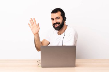 Tele pazarlamacı beyaz adam kulaklıkla çalışıyor ve laptopuyla selamlıyor..