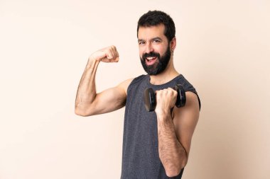 Beyaz sakallı sporcu arka planda ağırlık kaldırıyor. Güçlü bir hareket yapıyor.