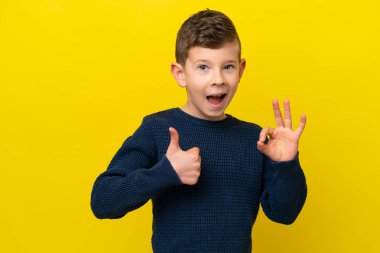 Sarı arka planda izole edilmiş küçük beyaz çocuk tamam işareti ve baş parmak işareti gösteriyor.