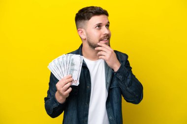 Kafkasyalı genç adam, sarı arka planda izole edilmiş bir şekilde çok para alıyor gülümserken yukarı bakıyor.