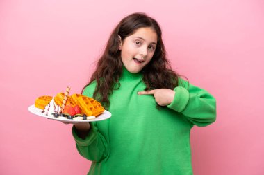 Pembe arka planda yüzünde sürpriz bir yüz ifadesiyle waffle tutan beyaz bir kız.