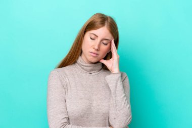 Genç kızıl saçlı kadın mavi arka planda baş ağrısı ile izole edilmiş.
