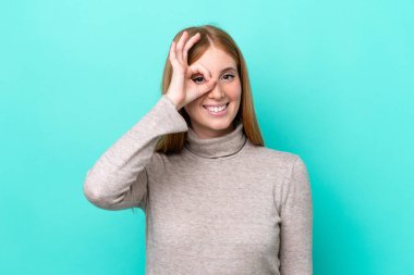 Mavi arka planda izole edilmiş genç kızıl saçlı kadın parmaklarıyla işaret veriyor.