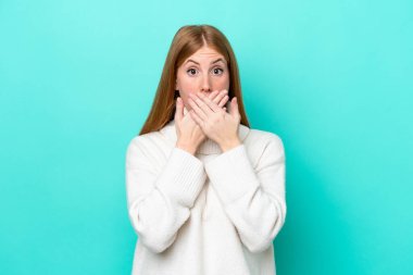 Mavi arka planda izole edilmiş genç kızıl saçlı kadın elleriyle ağzını kapatıyor.