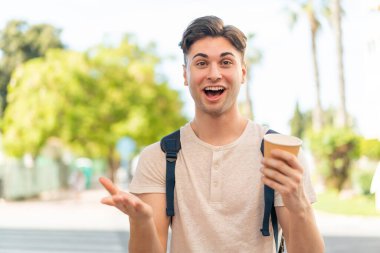 Genç yakışıklı bir adam açık havada kahve tutuyor ve yüzünde şok ifadesi var.