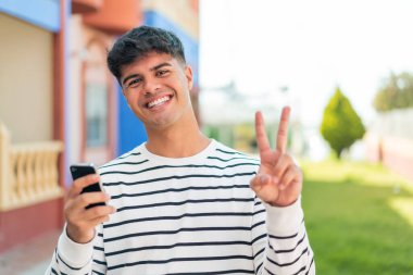 Genç İspanyol adam dışarıda cep telefonu kullanıyor. Gülümsüyor ve zafer işareti gösteriyor.