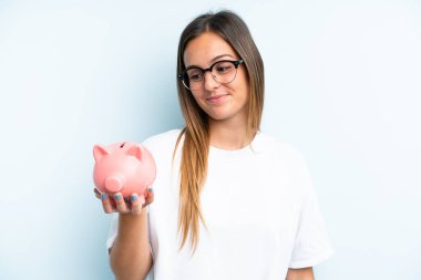 Elinde kumbara tutan beyaz bir kadın mavi arka planda mutlu bir ifadeyle izole edilmiş.