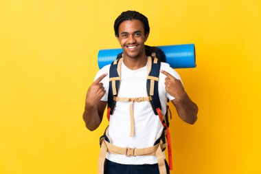 Sarı arka planda izole edilmiş büyük bir sırt çantalı genç dağcı, baş parmak hareketi yapıyor.