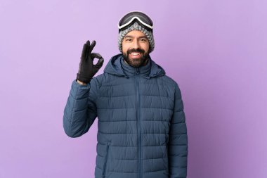 İzole edilmiş mor arka planda snowboard gözlüklü kayakçı parmaklarıyla tamam işareti gösteriyor.
