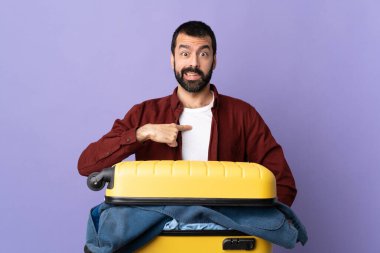 Bir bavul dolusu elbisesi olan beyaz bir adam kendini gösteren izole edilmiş mor bir arka plan.