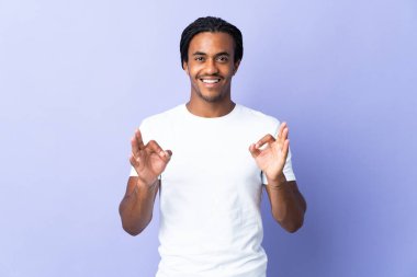 Örgülü genç Afrikalı Amerikalı adam mor arka planda izole edilmiş. İki eliyle tamam işareti gösteriyor.