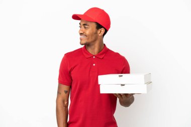 Afro-Amerikalı pizza teslimatçısı izole edilmiş beyaz duvarın üzerinden pizza kutuları alıp yan tarafa bakıyor ve gülümsüyor.
