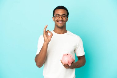 Afro-Amerikalı yakışıklı bir adam kumbarasını izole edilmiş mavi arka planda tutuyor. Parmaklarıyla işaret veriyor.