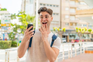 Dışarıda cep telefonu kullanan yakışıklı bir genç. Şaşırmış ve şok olmuş bir yüz ifadesi var.