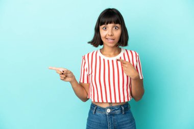 Mavi arka planda izole edilmiş genç melez kadın şaşırmış ve işaret ediyor.
