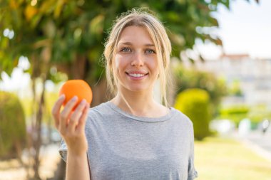 Açık havada elinde portakal tutan genç sarışın kadın çok gülümsüyor.