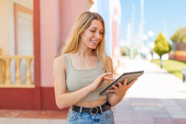 Açık havada genç sarışın kadın tablet ekranına mutlu bir ifadeyle dokunuyor.