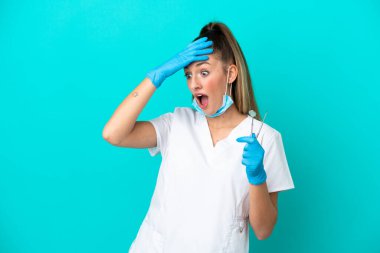 Beyaz dişçi kadın, elinde mavi arka planda izole edilmiş aletler tutuyor. Yan tarafa bakarken sürpriz jest yapıyor.