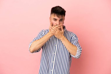 Pembe arka planda izole edilmiş genç beyaz adam elleriyle ağzını kapatıyor.